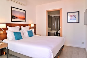 A hotel room with a large bed, a wooden headboard, a nightstand, a framed picture of a sunset, a bathroom with a shower, and a framed picture on the wall. - Hotel La Roya, an SLH Hotel (Saint-Florent)