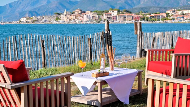 Una terraza frente al mar con muebles de madera, una mesa con una botella y vasos, y vistas a un pueblo costero y montañas.