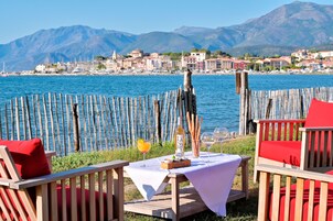 Una terraza frente al mar con muebles de madera, una mesa con una botella y vasos, y vistas a un pueblo costero y montañas.