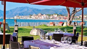 Un área de comedor al aire libre con mesas dispuestas para dos, con vistas a un cuerpo de agua y un paisaje montañoso.