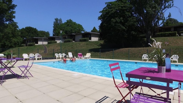 A swimming pool with lounge chairs and tables, surrounded by a landscaped area with trees and a hill. - Vvf Villages Parent (Parent)