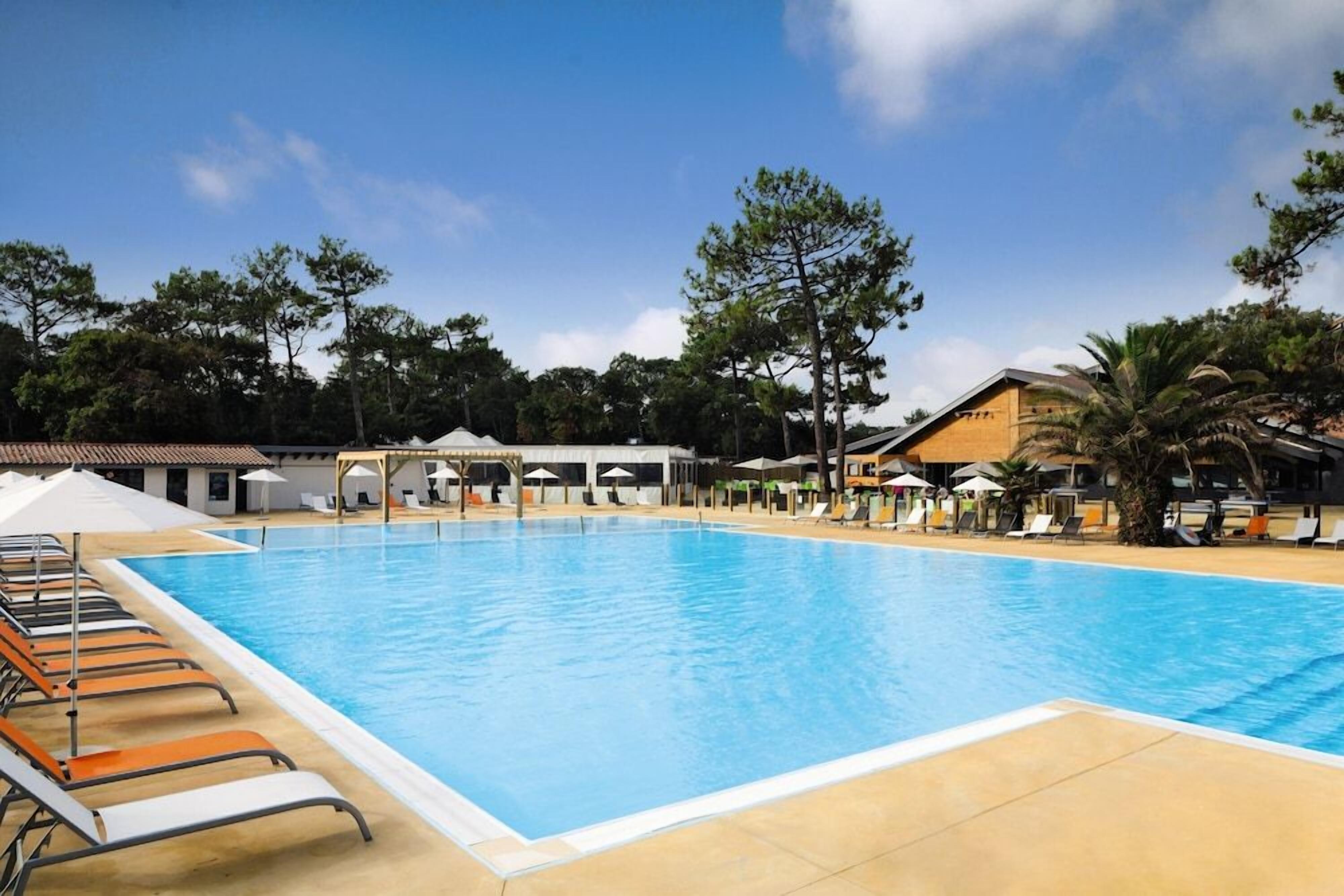 A large swimming pool with lounge chairs and umbrellas, surrounded by trees and buildings.