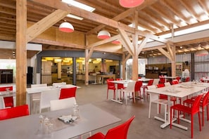Un comedor amplio con vigas de madera altas y sillas rojas. Las mesas están dispuestas con manteles blancos y cristalería.