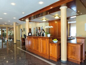 A hotel reception area with a wooden reception desk, a white vase with flowers, and a marble floor. - Victor S Residenz Teistungenburg (Teistungen)