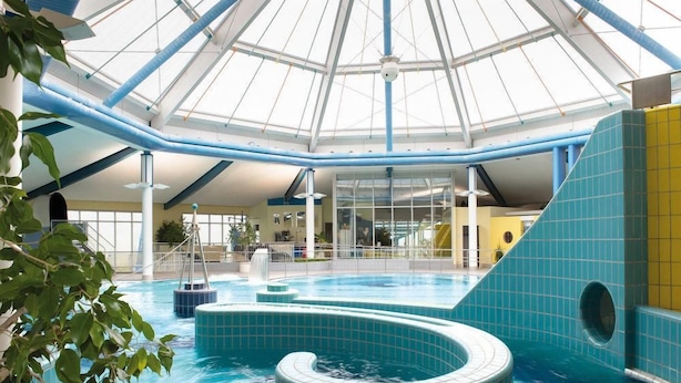 Una piscina interior con techo de vidrio, azulejos azules y un elemento central de agua.