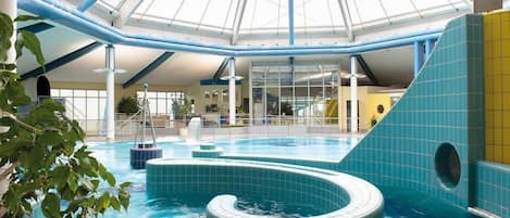 An indoor swimming pool with a glass ceiling, blue tiles, and a central water feature.