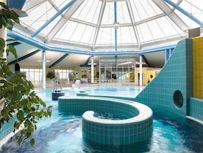 Una piscina interior con techo de vidrio, azulejos azules y un elemento central de agua.