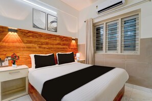 Ein Hotelzimmer mit einem Holzkopfteil, einem Bett mit schwarz-weißer Bettwäsche, zwei Nachttischlampen, einem Nachttisch mit Blumenvase und einem Fenster mit Jalousien.