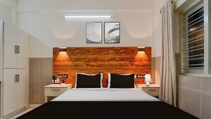 Une chambre d’hôtel comprenant un lit, deux tables de chevet, une petite table, une lampe fixée au mur, une fenêtre avec des rideaux et une tête de lit en bois.