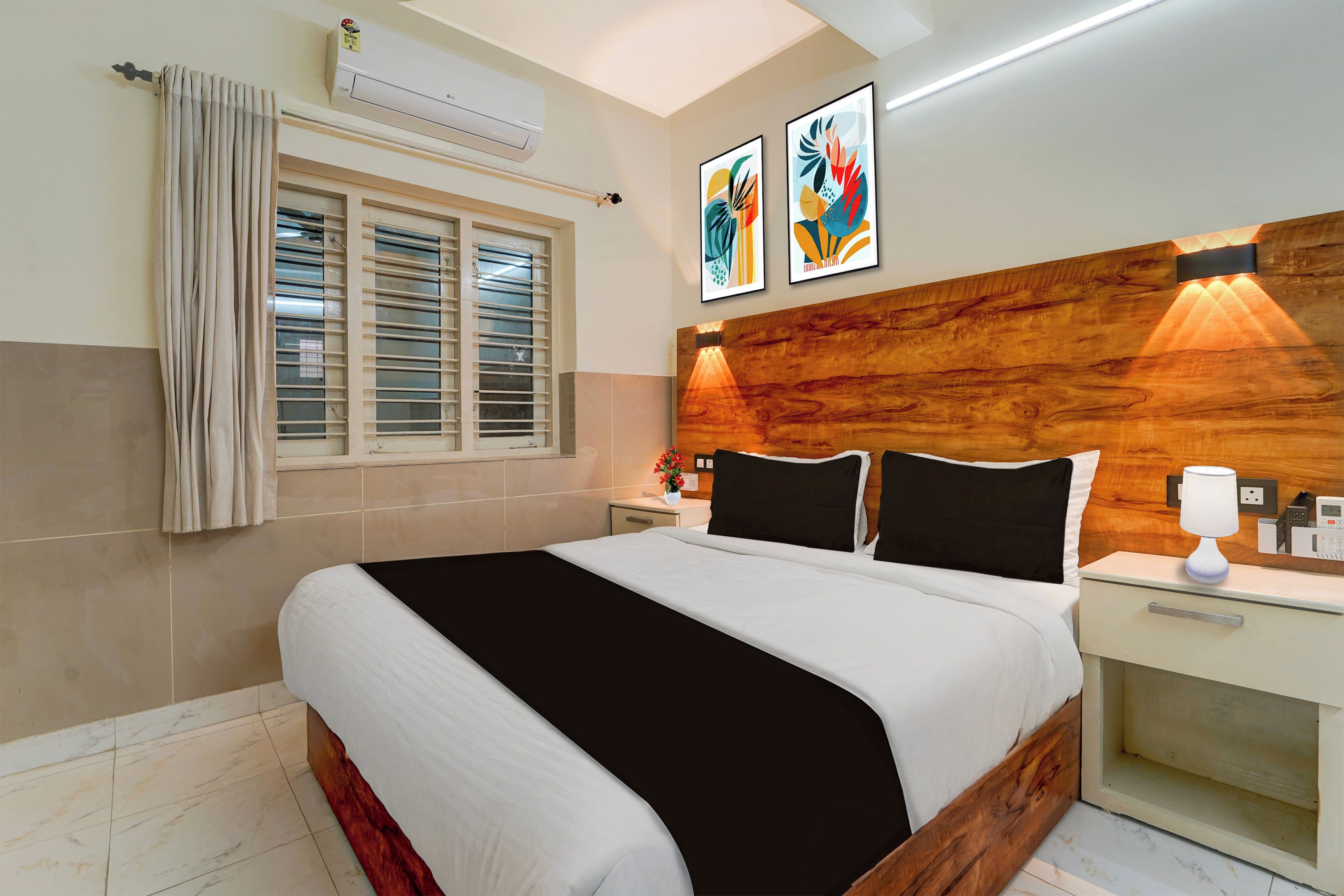 A hotel room with a bed, a nightstand, a bedside table, a wall-mounted air conditioner, a window with blinds, and two framed artworks on the wall.