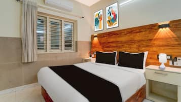A hotel room with a bed, a nightstand, a bedside table, a wall-mounted air conditioner, a window with blinds, and two framed artworks on the wall.