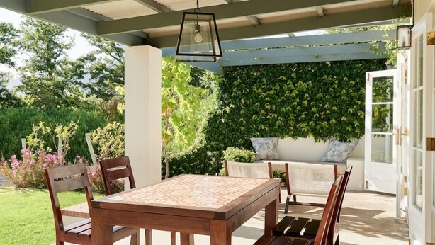 En udendørs terrasse med et træbord og stole, en hængende lampe og en væg dækket af grøn vegetation.