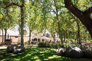 Un área de estar al aire libre con mesas y sillas bajo los árboles, un edificio al fondo y un primer plano con césped.