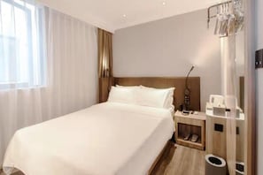 Une chambre d’hôtel avec un grand lit, une table de chevet, une commode, une lampe, un porte-chaussures et une poubelle.