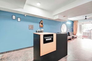 Un mostrador de recepción con la etiqueta 'TOUMHOUSE Company Serviced', una pared azul, cuadros enmarcados, un pequeño santuario y una zona de estar con un espejo redondo.