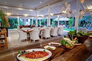 A restaurant with a tropical theme, featuring a large fish dish, a bowl of vegetables, and a view of the garden. - Villa Sipo (Keroboken Kelod)