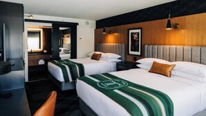 Habitación de hotel con dos camas, un escritorio, una silla y un baño visible a través de una puerta abierta.