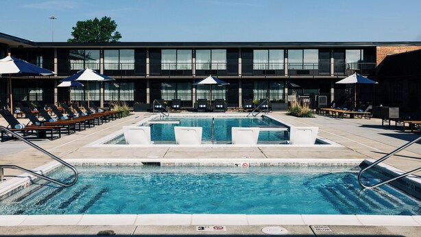Un área de piscina con sillones de descanso y sombrillas, un edificio con ventanas amplias y un cielo despejado.