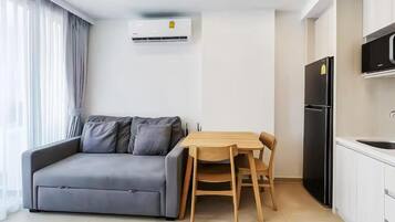 A modern living space with a grey sofa, wooden dining table, and chairs, a black refrigerator, and a white air conditioning unit.