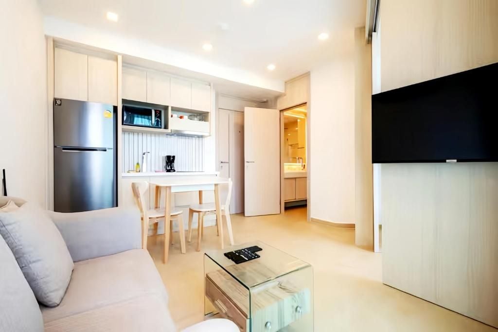 A modern kitchen with white cabinetry, a built-in microwave, and a coffee machine. There is a glass coffee table in the living area.
