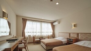 Una habitación de hotel con escritorio, dos camas, una silla y un amplio ventanal con cortinas.
