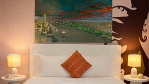 Een hotelkamer met een bed, twee bedlampen, een geborduurd plaid, een kussen en een schilderij aan de muur.
