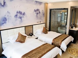 Habitación de hotel con dos camas, un cuadro grande en la pared, cabina de ducha con paredes de vidrio y una mesita de noche con lámpara.