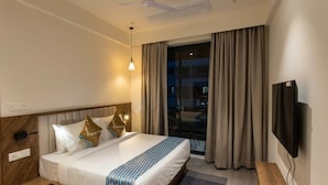 Una habitación de hotel con cama, televisión, ventilador de techo y ventana con cortinas.