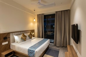 A hotel room with a bed, a TV, a ceiling fan, and a window with curtains. - Townhouse Sector 143B Noida (Noida)