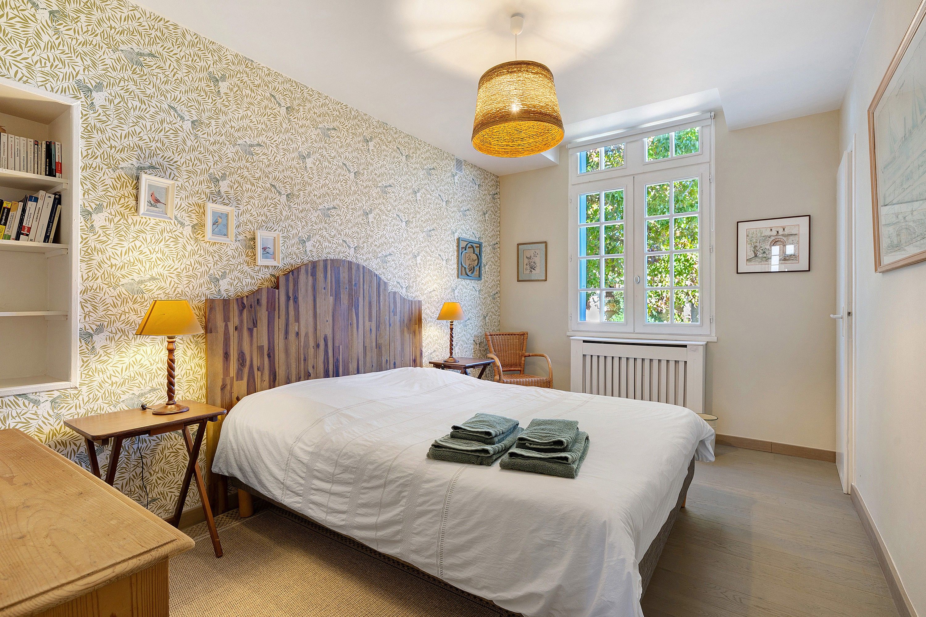 Une chambre à coucher avec un grand lit, une tête de lit en bois, une fenêtre donnant sur un beau paysage, une bibliothèque et une lampe.