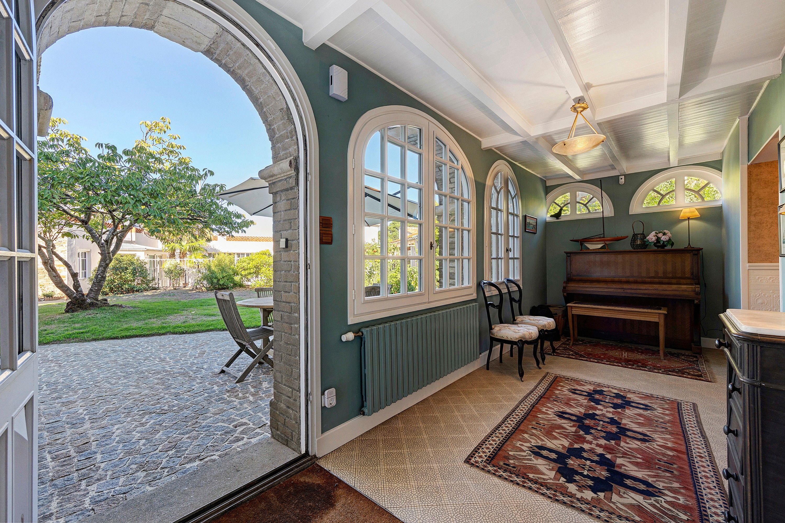 Une pièce dotée d’une arche en pierre menant à un patio, avec un piano et un coin repas comprenant des chaises.