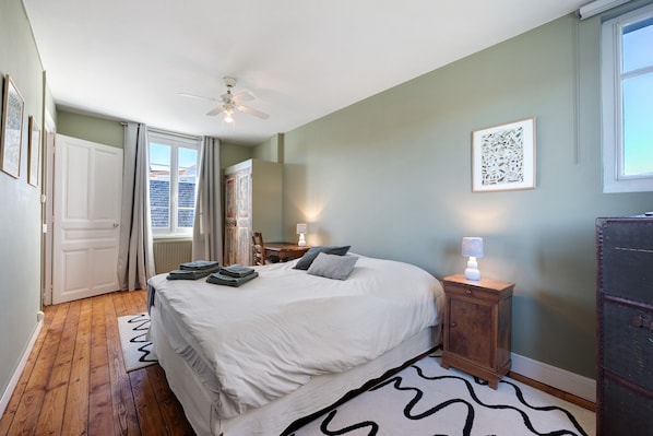 A bedroom with a bed, a bedside table with a lamp, a ceiling fan, and a window with curtains. - Villa Serpentine Guest Room – Family Suite (Les Sables-d'Olonne)