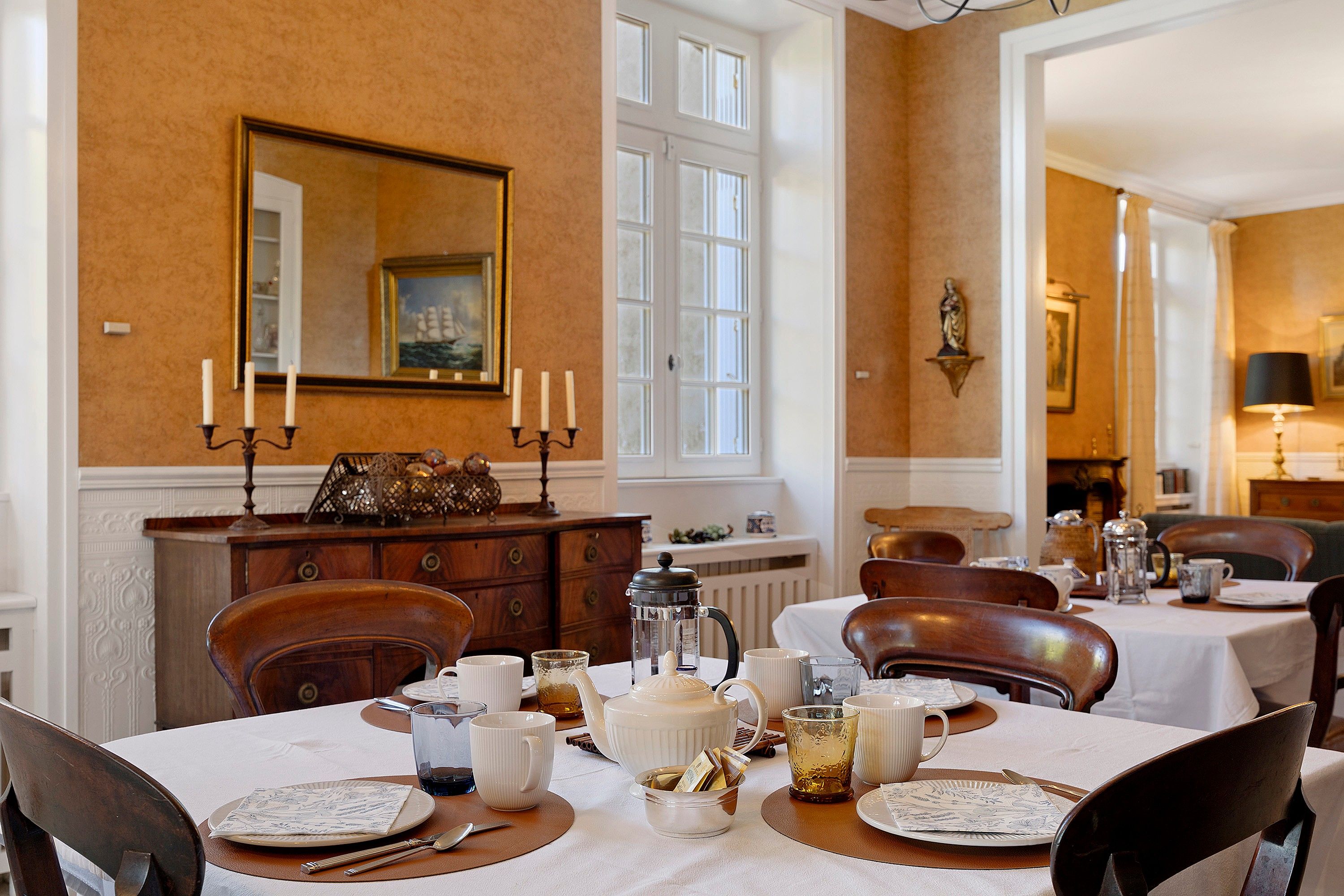 A dining area with a table set for four, a mirror on the wall, and a statue above the fireplace.