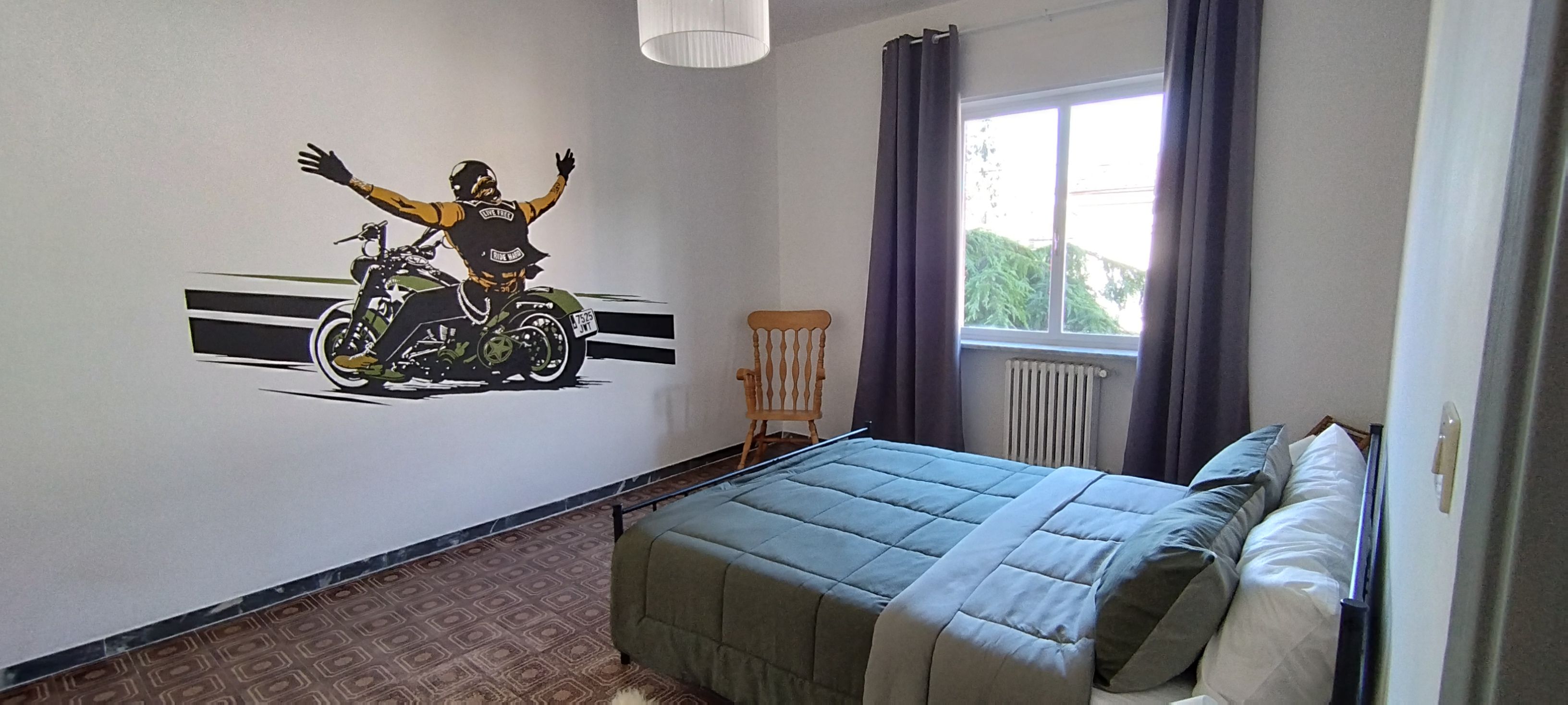 Une chambre à coucher avec un lit, une fenêtre, un radiateur, une chaise et une fresque murale représentant une moto.