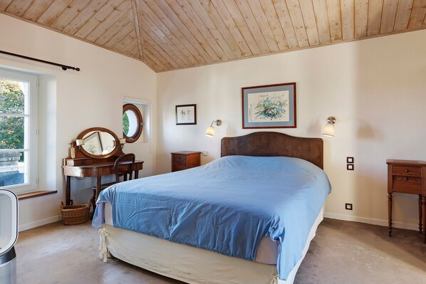 A bedroom with a large bed, a wooden desk with a mirror, a dresser, and a window with a view of greenery. - Villa Serpentine Guest Room – Parental Suite (Les Sables-d'Olonne)