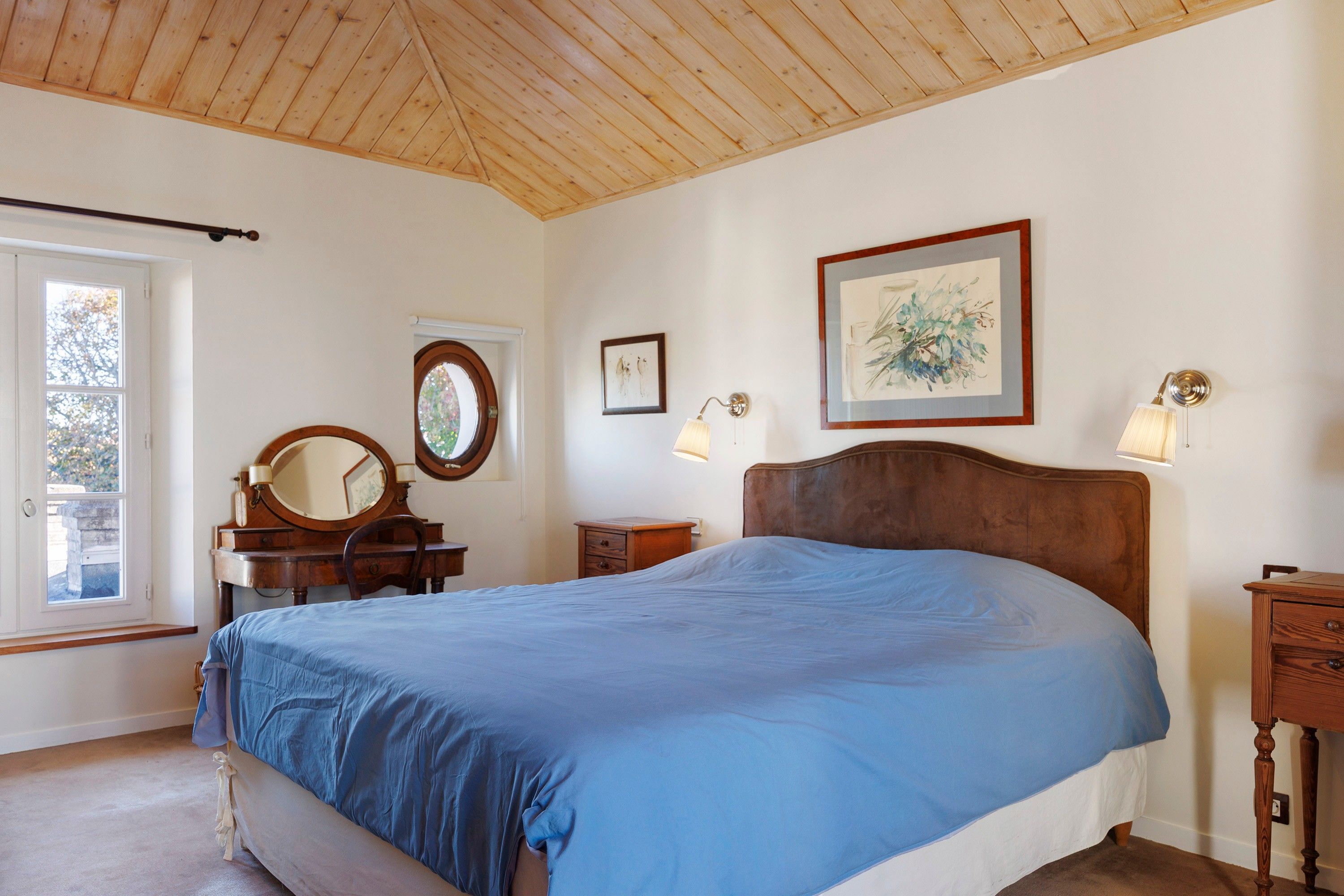 Een slaapkamer met een houten plafond, een bed met een blauwe plaid, een houten dressoir, een ronde spiegel, een lijst met een foto aan de muur en een raam met uitzicht op bomen.