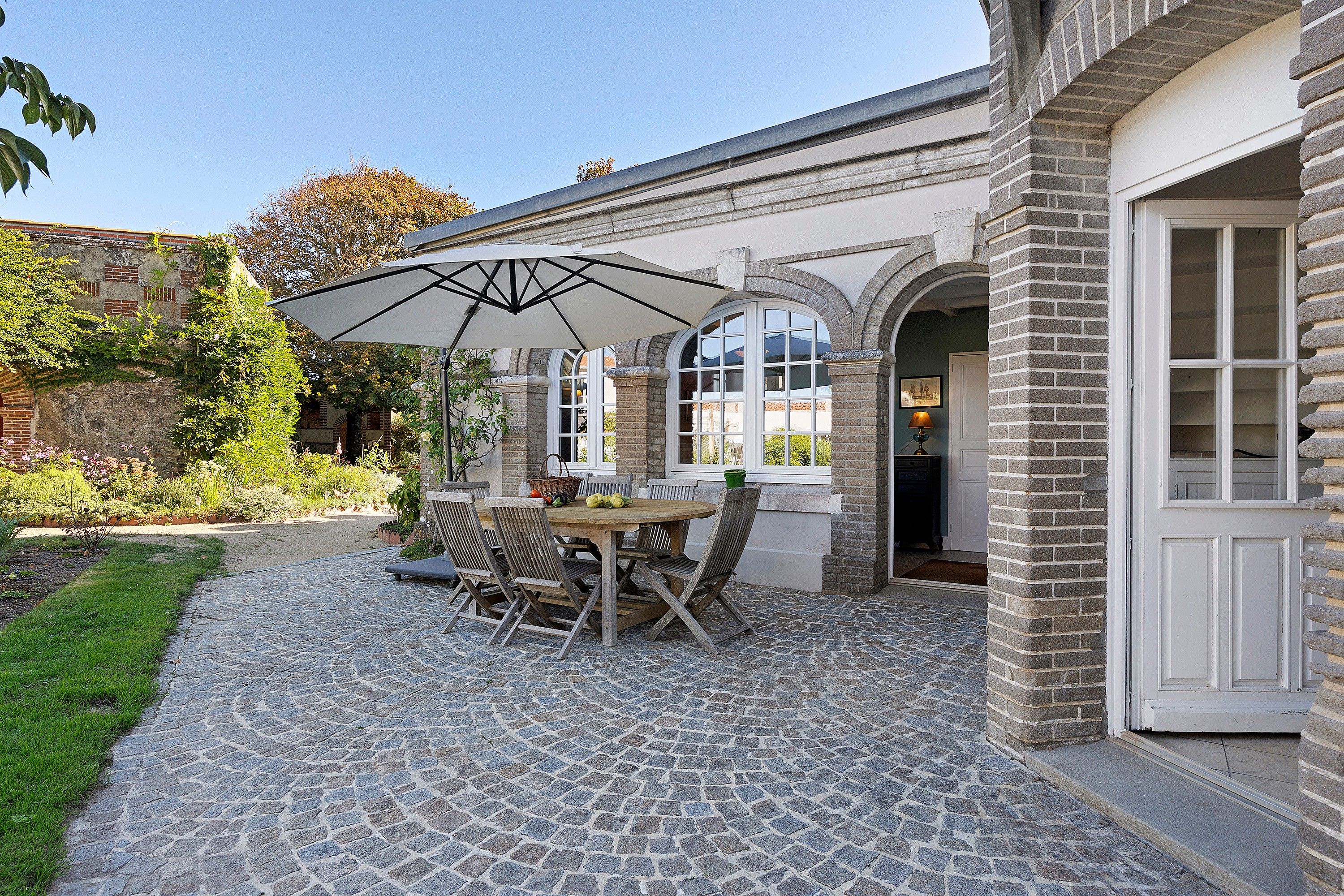 Un espace patio doté d’un sol carrelé en pierre, d’une table en bois avec des chaises et d’un grand parasol offrant de l’ombre.