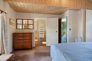 A bedroom with a wooden ceiling, a large bed, a dresser, a radiator, and a bathroom visible through an open door.