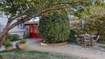 Un patio con un sendero de grava que conduce a una puerta roja, rodeado de follaje y una pequeña mesa con asientos al aire libre.