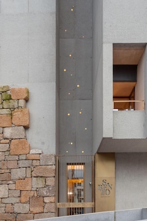 Un bâtiment avec un mur en pierre, une façade en béton et une porte sur laquelle est apposée une enseigne.