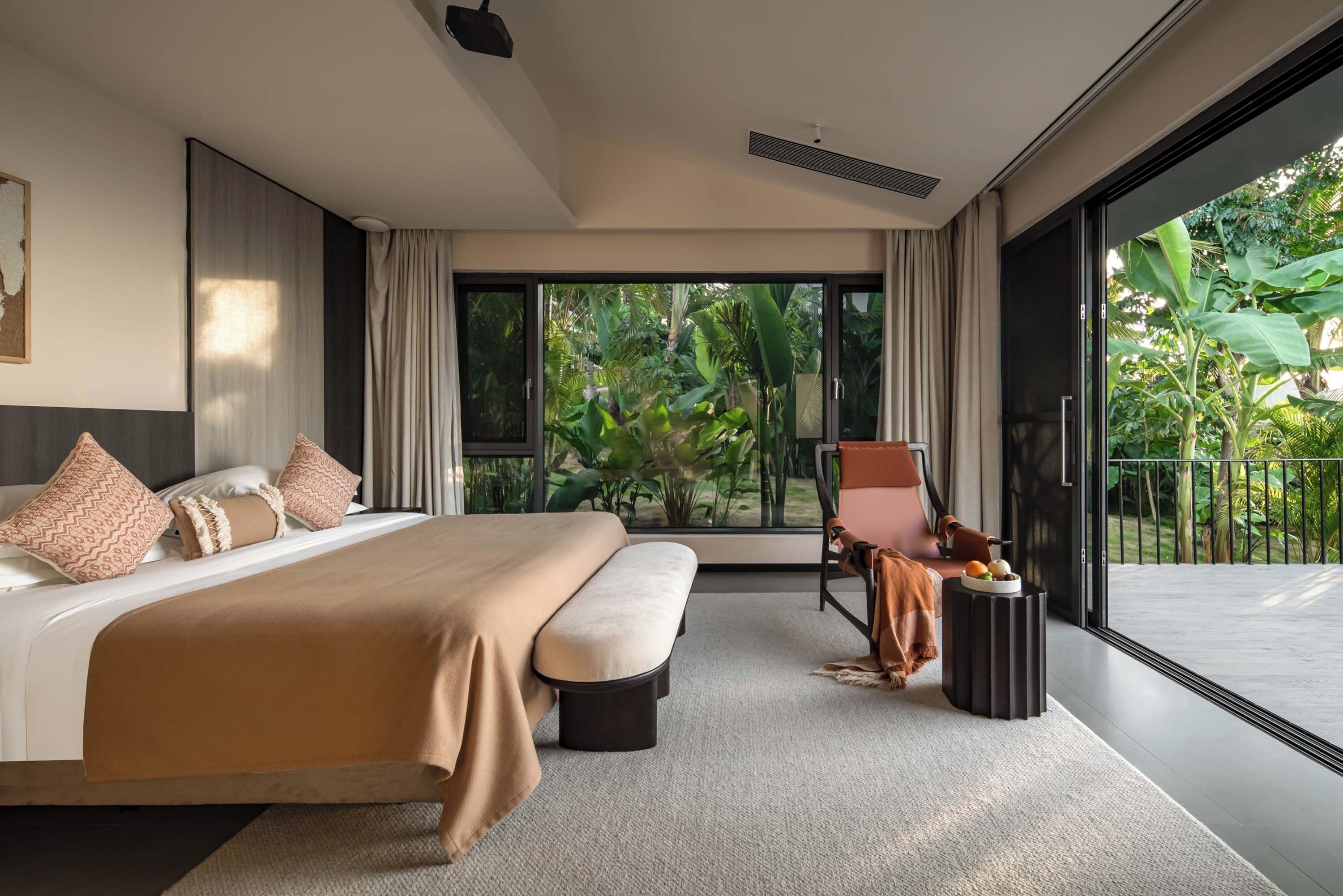 A modern bedroom with a large bed, a seating area with a chair, and a view of lush greenery outside.