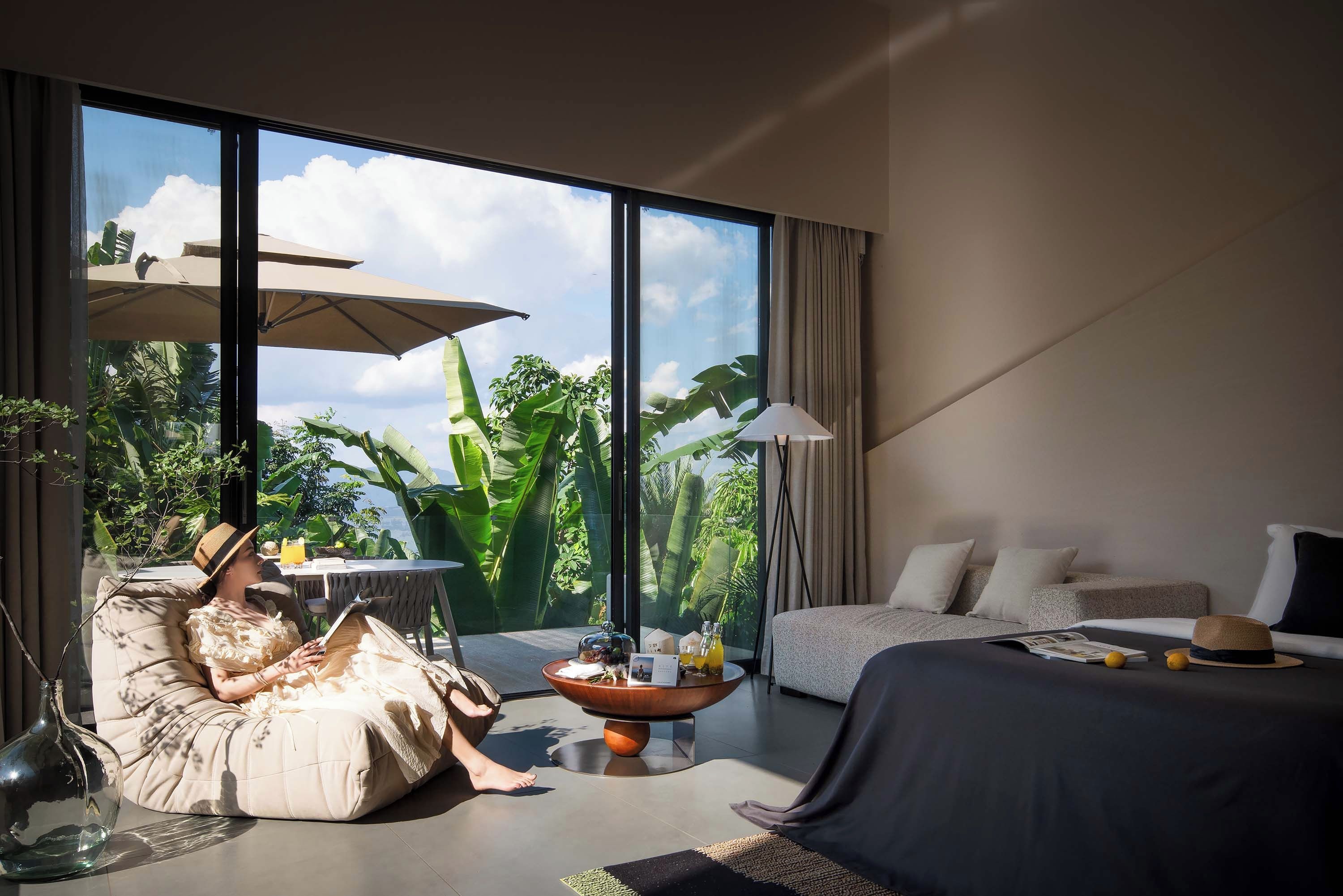 A modern hotel room with a large window, a sofa, a bed, and a dining area with a table and chairs.