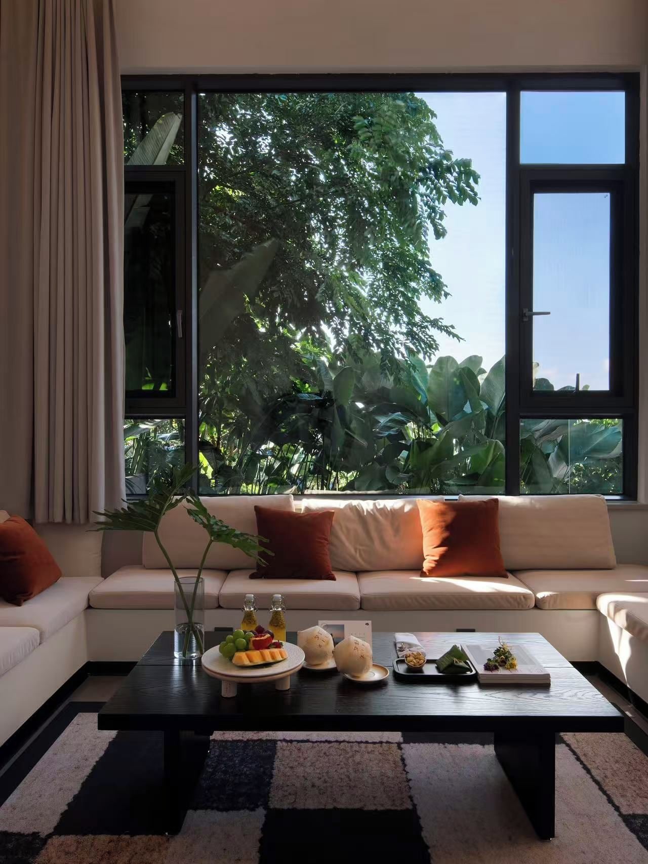 A modern living room with a large window, a sofa, a coffee table with fruit and drinks, and a view of greenery outside.
