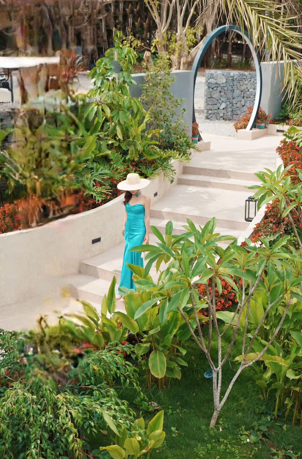Una mujer con un vestido azul sube unas escaleras exteriores rodeada de exuberante vegetación y plantas.