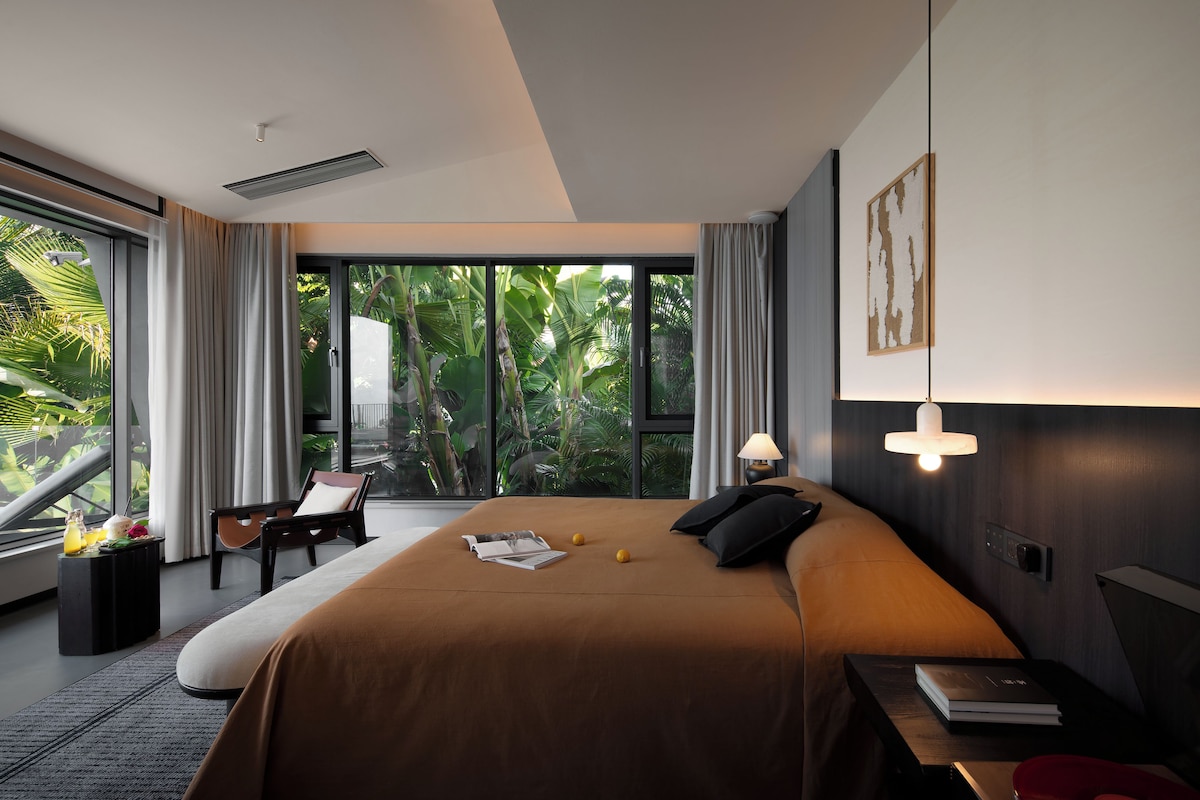 Een moderne hotelkamer met een groot bed, een zithoek met een fauteuil, een tafeltje en een groot raam met uitzicht op groen.