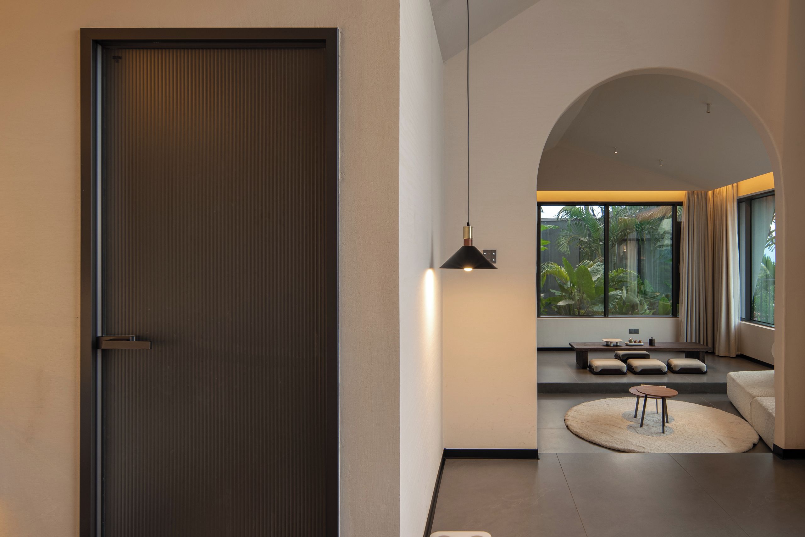 A modern interior with a sliding door, a hanging pendant light, a seating area with cushions, and a view through a large window.
