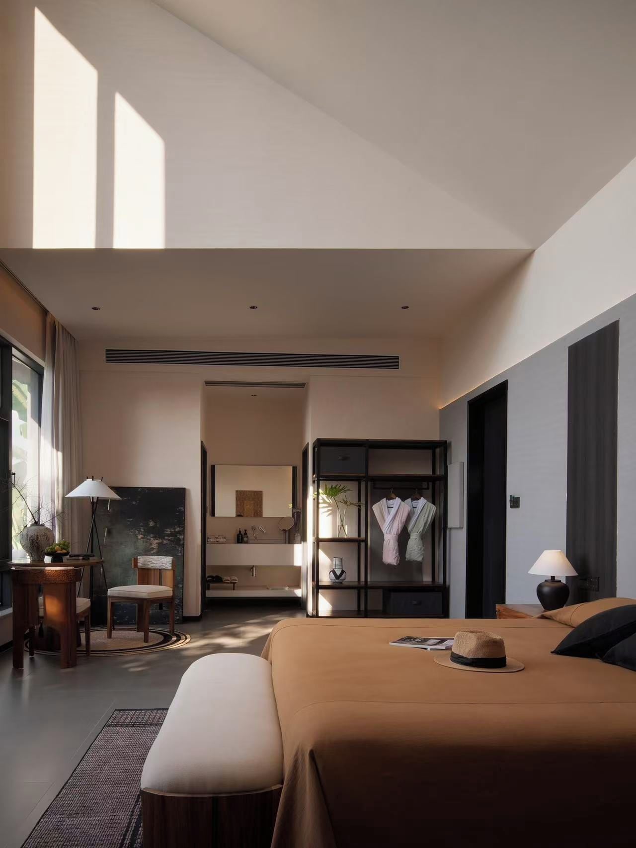 A modern hotel room with a large bed, a sitting area with a chair and table, a vanity with a mirror, and a walk-in closet with hanging robes.