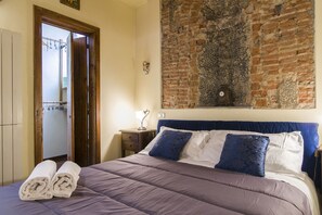 Een slaapkamer met een bed, een houten deur, een bakstenen muur en een klein tafeltje met een lamp.