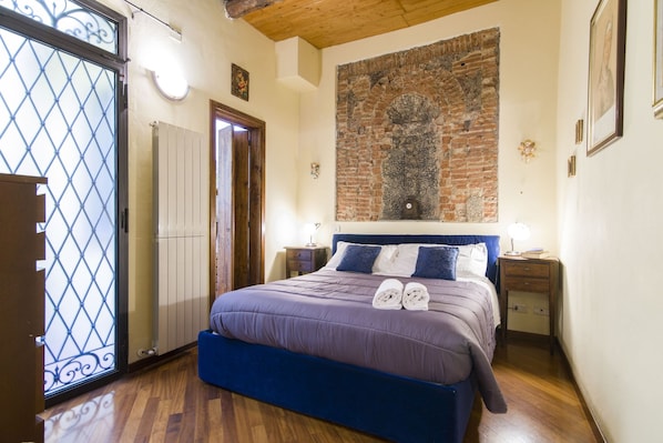 Een slaapkamer met een bed, een houten vloer, een bakstenen muur en een raam met een metalen traliewerk.