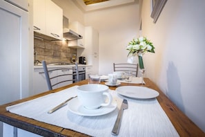 Una mesa de comedor dispuesta con platos blancos, tazas, cubiertos, un florero con flores blancas y una silla de madera.